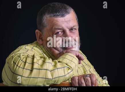 Bel ritratto di un uomo anziano sorridente in camicia gialla appoggiò il gomito su uno schienale di sedia contro sfondo nero Foto Stock