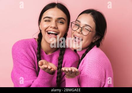 Immagine di due ragazze adolescenti felici con trecce in abiti casual sorridente e soffiare aria bacio a voi isolato su sfondo rosa Foto Stock