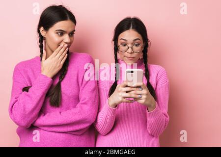 Immagine di giovane ragazza adolescente in piedi da felice amico femminile e peeking al suo cellulare isolato su sfondo rosa Foto Stock