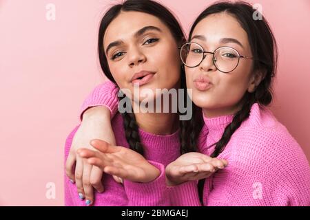 Immagine di due ragazze adolescenti felici con trecce in abiti casual sorridente e soffiare aria bacio a voi isolato su sfondo rosa Foto Stock