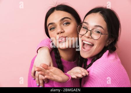 Immagine di due ragazze adolescenti felici con trecce in abiti casual sorridente e soffiare aria bacio a voi isolato su sfondo rosa Foto Stock