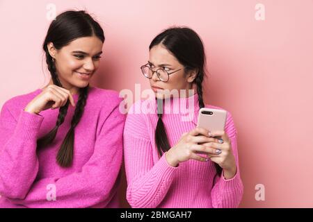 Immagine di giovane ragazza adolescente in piedi da felice amico femminile e peeking al suo cellulare isolato su sfondo rosa Foto Stock