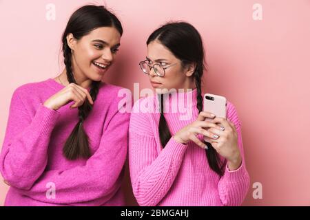 Immagine di giovane ragazza adolescente in piedi da felice amico femminile e peeking al suo cellulare isolato su sfondo rosa Foto Stock