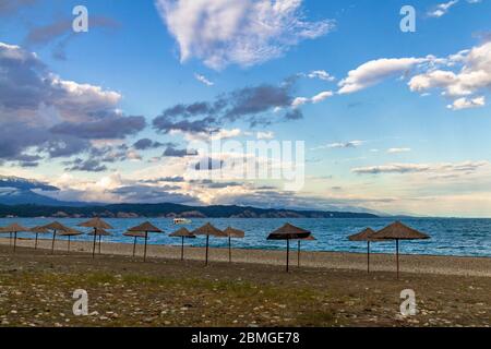 Ombrelloni da spiaggia di paglia sulla costa vuota del Mar Nero in Abkhazia, Pitsunda al tramonto. Spiaggia di ghiaia, montagne, mare, cielo con nuvole Foto Stock