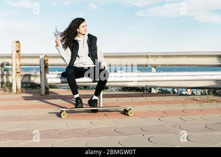 Adolescente femmina seduta su una panchina con i piedi sulla longboard di fronte al mare Foto Stock