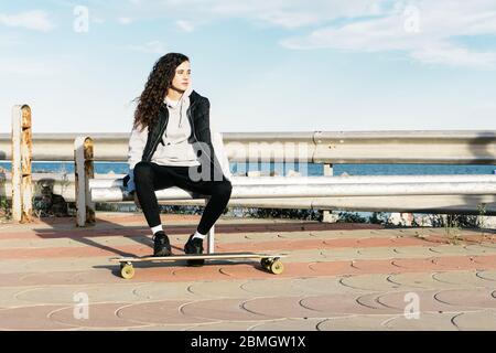 Adolescente femmina seduta su una panchina con i piedi sulla longboard di fronte al mare Foto Stock