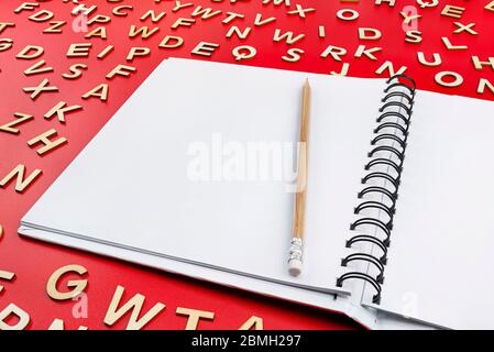 Un notebook aperto con pagine bianche e una matita di legno adagiata su uno sfondo rosso con lettere sparse in legno. Foto Stock