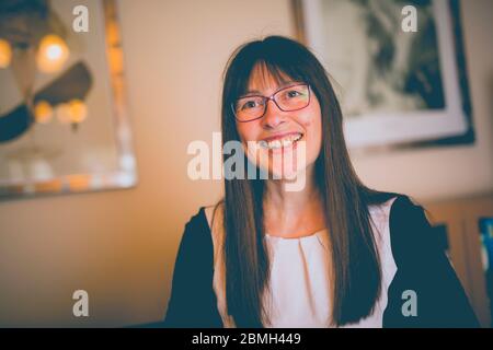 Vista frontale in primo piano di una donna britannica in occhiali sorridenti, signora isolata al chiuso, soggiorno nel Regno Unito. Foto Stock