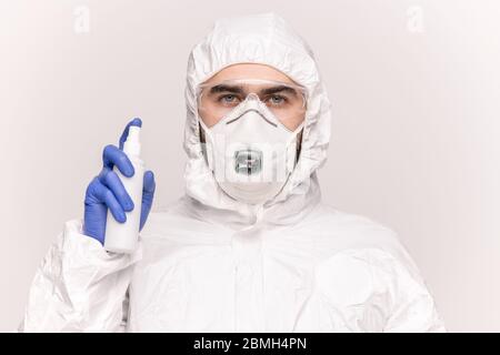 Bel giovane uomo in tute protettive, respiratore, guanti e occhiali spruzzando disinfettante intorno a sé in isolamento Foto Stock