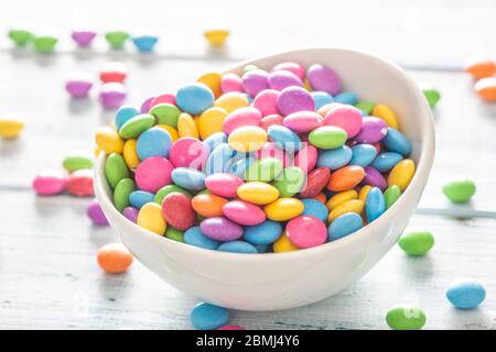 Primo piano di una ciotola bianca piena di smarties con molti posti a caso intorno. Foto Stock