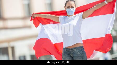 Giovane donna in maschera facciale tiene bandiera austriaca sulla strada. Foto Stock