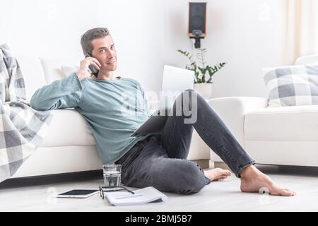 Abiti casual indossando l'uomo si siede sul pavimento dell'appartamento, chiamando su un telefono cellulare con un notebook davanti a lui. Foto Stock