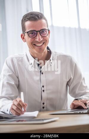 Il giovane uomo d'affari che indossa gli occhiali sorride alla macchina fotografica mentre si siede alla sua scrivania. Foto Stock