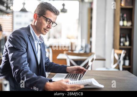 Giovane uomo d'affari in una giacca e bicchieri legge in una caffetteria con un computer su un tavolo davanti a lui. Foto Stock
