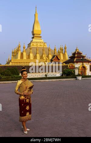 Bella giovane donna felice vestita con abiti tradizionali etnici del Laos davanti a Pha That Luang, o grande Stupa a Vientiane, Laos City Center Foto Stock