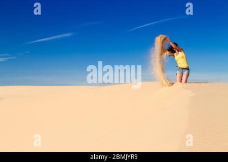 Donna siede sulla sabbia su una cresta nel deserto, getta sabbia nell'aria. Foto Stock