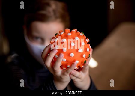 Il bambino che indossa una maschera medica tiene in mano un giocattolo di gomma. Periodo di isolamento a casa, protezione durante l'epidemia coronavirus. Il giocattolo assomiglia a un virus.Model Foto Stock