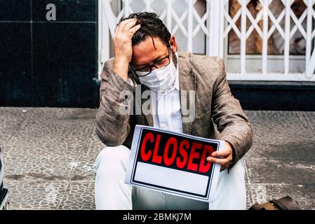 Uomo d'affari triste e disperato sedersi fuori del suo negozio chiuso per coronavirus blocco economia crisi di affari - persone con maschera di protezione per vi Foto Stock