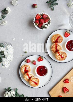 Frittelle di formaggio ucraino fatte in casa Syrniki con marmellata di fragole rosse e frutti vicino a fiori bianchi sulla vista da tavolo in stoffa Foto Stock