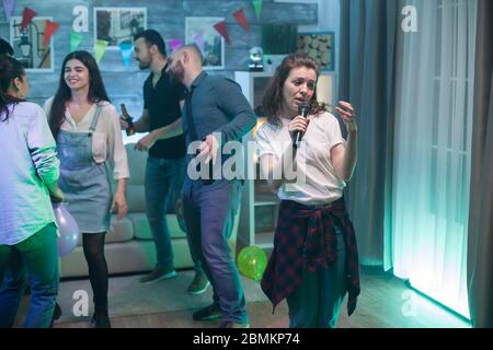 Bella giovane donna in una t-shirt bianca facendo karaoke ad una festa con gruppo di amici. Foto Stock