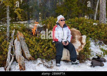 Donne anziane attive che riposano seduti su una pelle di renne con un fuoco da campeggio e la foresta sullo sfondo, foto di Mellansel Vasternorrland, Sverige. Foto Stock