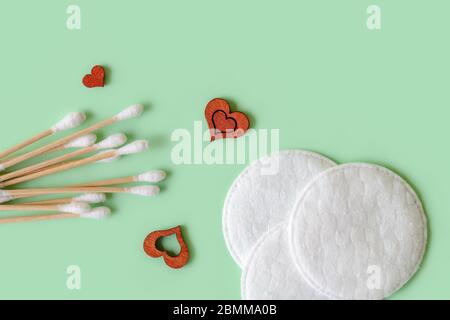Bastone di legno di gemme di cotone, tamponi di cotone e pochi cuori di legno piccoli su uno sfondo di menta pastello. Concetto di materiali eco-compatibili per la cura del viso e del corpo. Foto Stock