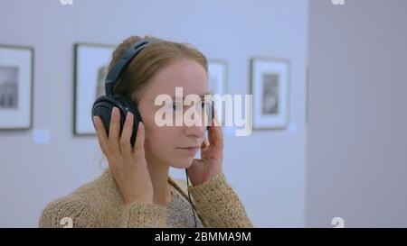 Donna che guarda la esposizione, utilizzando le cuffie e ascolto audio guida in una moderna galleria fotografica. Istruzione e concetto di intrattenimento Foto Stock