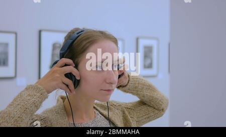 Donna che guarda la esposizione, utilizzando le cuffie e ascolto audio guida in una moderna galleria fotografica. Istruzione e concetto di intrattenimento Foto Stock