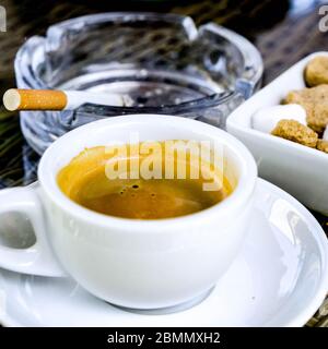 Tazza di caffè espresso forte con un bicchiere di zucchero e una sigaretta illuminata in un posacenere di vetro, senza persone Foto Stock