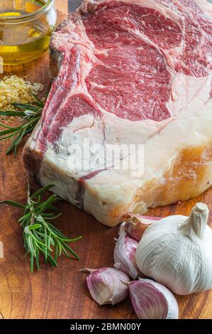 Bistecca di vaca rubia gallega invecchiata, pronta per essere grigliata con aglio, rosmarino, scaglie di sale e olio extravergine di oliva e coltello da cucina su tavola di legno Foto Stock