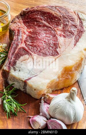 Bistecca di vaca rubia gallega invecchiata, pronta per essere grigliata con aglio, rosmarino, scaglie di sale e olio extravergine di oliva e coltello da cucina su tavola di legno Foto Stock