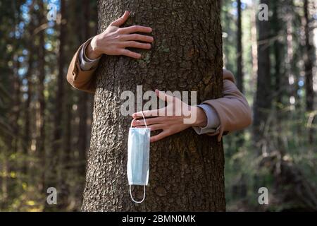 Primo piano delle mani che abbracciano un albero e toglie la maschera medica protettiva, lo tiene sulle sue dita, gode di vita, aria pulita fresca nella foresta dopo Co Foto Stock