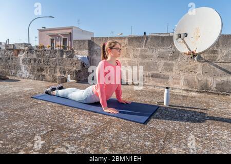 Giovane ragazza sta facendo verso l'alto Dog posa, Urdhva Mukha Svanasana sul tetto della sua casa durante la quarantena di coronavirus. Training di yoga femminile per Foto Stock