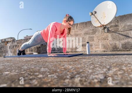 Giovane ragazza sta facendo asse sul tappeto sul tetto della sua casa durante la quarantena di coronavirus. Allenamento di yoga femminile per mantenersi in salute. allenamenti domestici Foto Stock