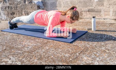 Giovane ragazza sta facendo asse sul tappeto sul tetto della sua casa durante la quarantena di coronavirus. Allenamento di yoga femminile per mantenersi in salute. allenamenti domestici Foto Stock