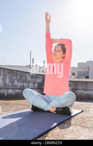 La ragazza giovane sta facendo lo yoga che si allunga sul tappeto sul tetto della sua casa durante il blocco di coronavirus. Allenamento di yoga femminile per rimanere in salute. Cura personale Foto Stock