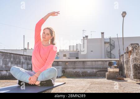 La ragazza giovane sta facendo lo yoga che si allunga sul tappeto sul tetto della sua casa durante il blocco di coronavirus. Allenamento di yoga femminile per rimanere in salute. Cura personale Foto Stock