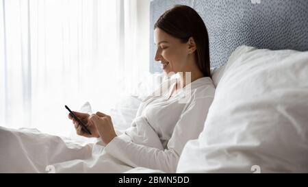 Bella donna che si trova a letto passare il tempo utilizzando lo smartphone Foto Stock