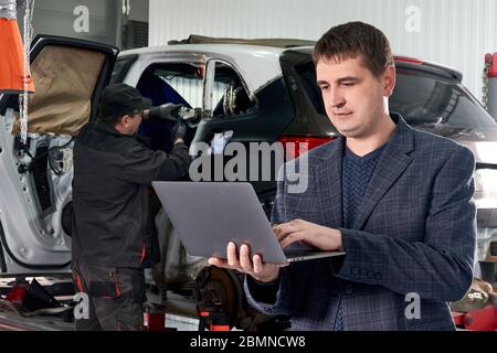 Il manager tiene il laptop mentre si trova vicino all'auto in un negozio di riparazione auto con un meccanico che ripara l'auto in background Foto Stock
