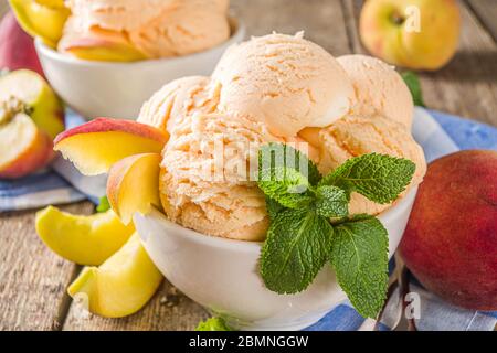 Gelato alla pesca dolce fatto in casa. Palle di gelato alla pesca in piccole ciotole, su sfondo di legno con pesche fresche e foglie di menta Foto Stock