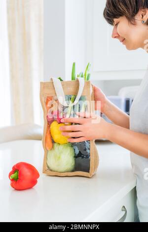 Donna che tiene riutilizzabile eco sacco tessuto sacco imballaggio con verdure biologiche fresche su tavolo da cucina bianco. Concetto di consegna a domicilio. Contadino locale lui Foto Stock