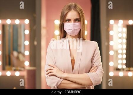 Vita su ritratto di bella giovane donna che indossa maschera rosa viso in piedi con le braccia incrociate mentre si posa in spogliatoio interno, spazio copia Foto Stock