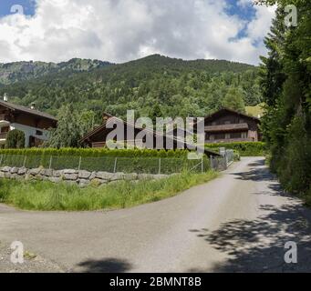 Chalet svizzeri classici lungo la strada locale rustica. Accogliente villaggio rurale Champery in Svizzera. Cielo blu brillante e nuvole bianche e morbide su picchi rocciosi. Foto Stock