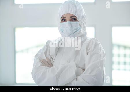 Operatore medico in indumenti protettivi e maschera medica in piedi con le braccia piegate Foto Stock