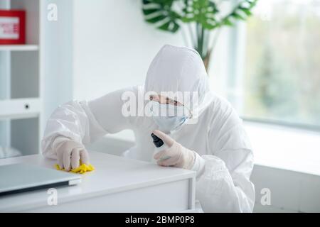 Operatore medico in indumenti protettivi e maschera medica spruzza disinfettante sul tavolo Foto Stock