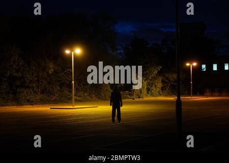 Una misteriosa figura con cappuccio, in piedi con la schiena alla macchina fotografica sotto le luci di strada di notte Foto Stock