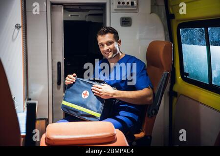 Un giovane paramedico maschile seduto in un'ambulanza, tenendo il suo kit medico e sorridendo alla macchina fotografica Foto Stock