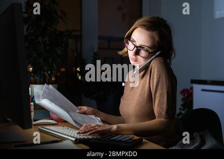 Donna in occhiali che utilizza la calcolatrice per calcolare la fattura, pianificare le spese, parlare sullo smartphone, lavorare sul PC desktop a tarda notte a casa offi Foto Stock