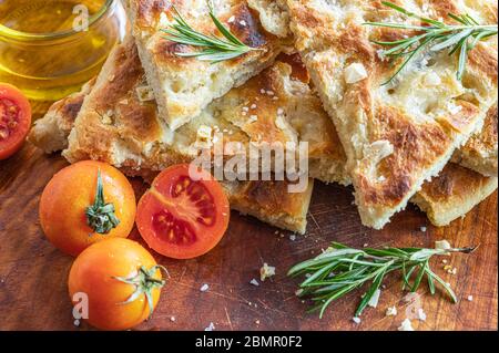 Focaccia ligure appena sfornata, tagliata a fette con pomodori, rosmarino, olio extravergine di oliva e scaglie di sale su una tavola di legno Foto Stock