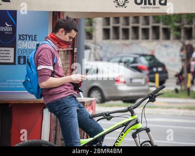 BELGRADO, SERBIA - 5 MAGGIO 2020: Giovane uomo in bicicletta che controlla le app sul suo telefono cellulare, uno smartphone, indossando una bandana come un dispositivo di protezione viso Foto Stock